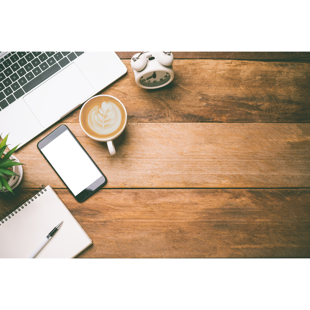 Overhead workspace view with laptop, coffee cup, notebook and phone on wooden table.