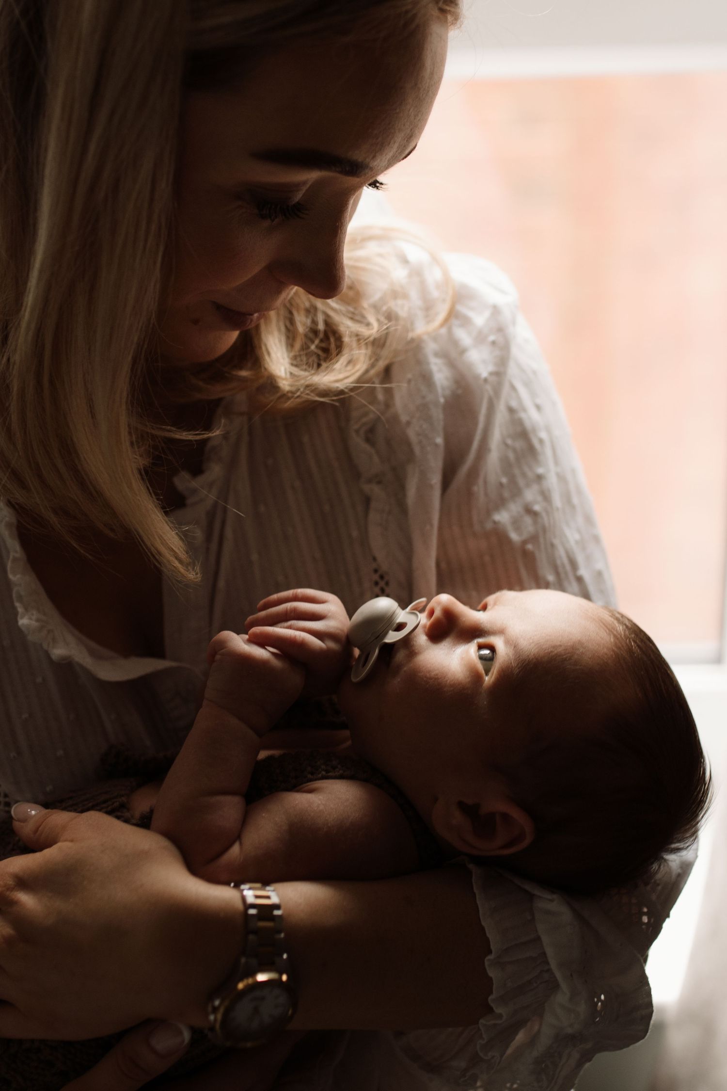 A blonde mother tenderly holds her newborn baby with a pacifier near a window in soft natural light.
