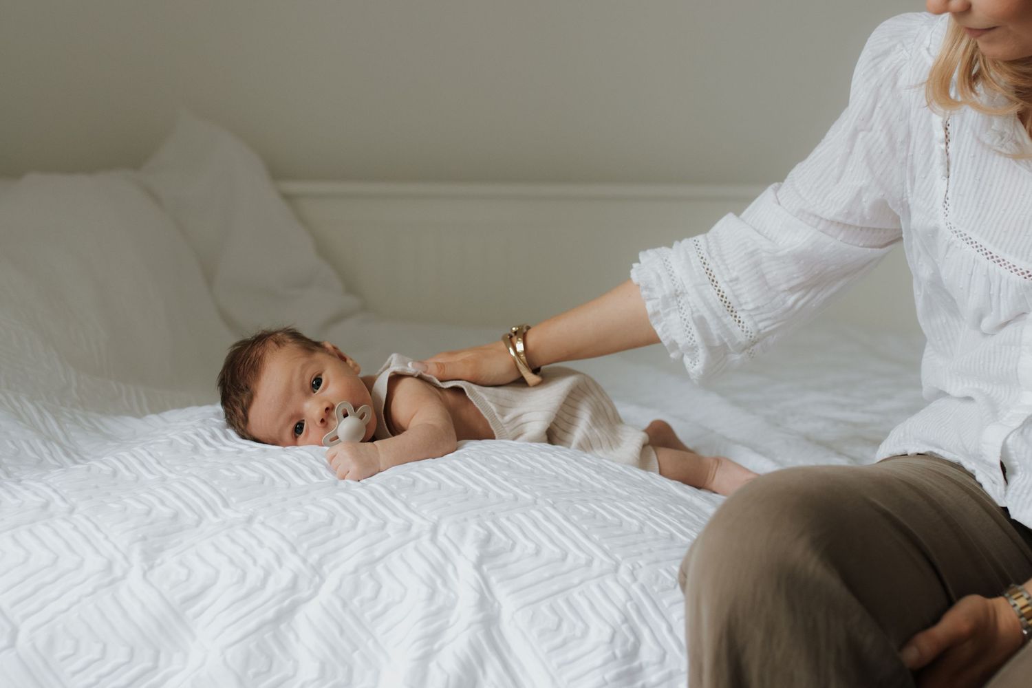 A mother in a white blouse gently holds her newborn baby lying on a white bed.