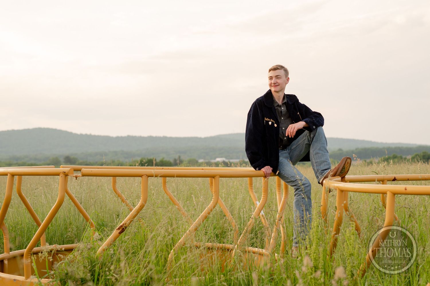 Casual pose on wooden fence with scenic rural landscape in background at sunset.