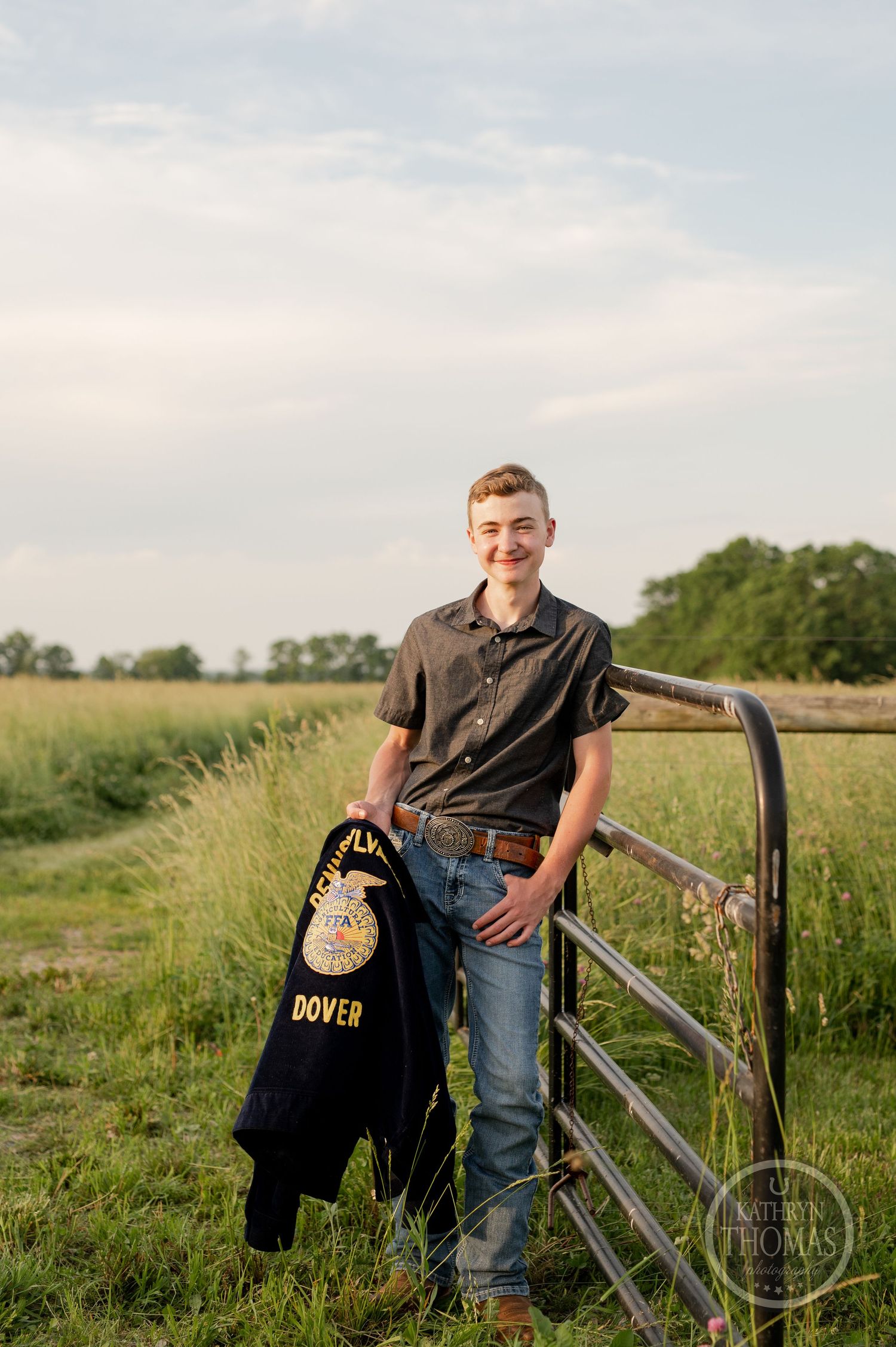 Student in casual attire poses with FFA jacket against rustic farm gate in grassy field.