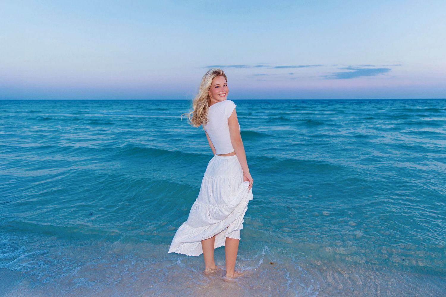 Person in white dress standing in shallow turquoise ocean water during blue hour.