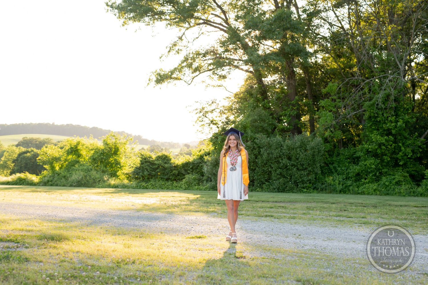 Sunset graduation photo with graduate standing on path near wooded area.
