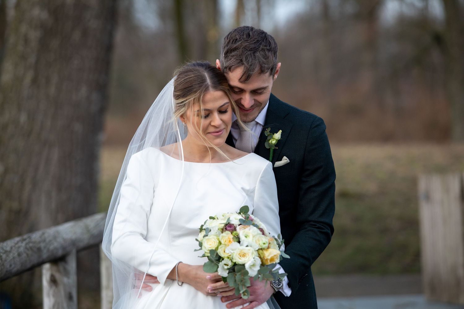 Romantic wedding moment as a couple shares a tender embrace while admiring the bridal bouquet in an outdoor setting.