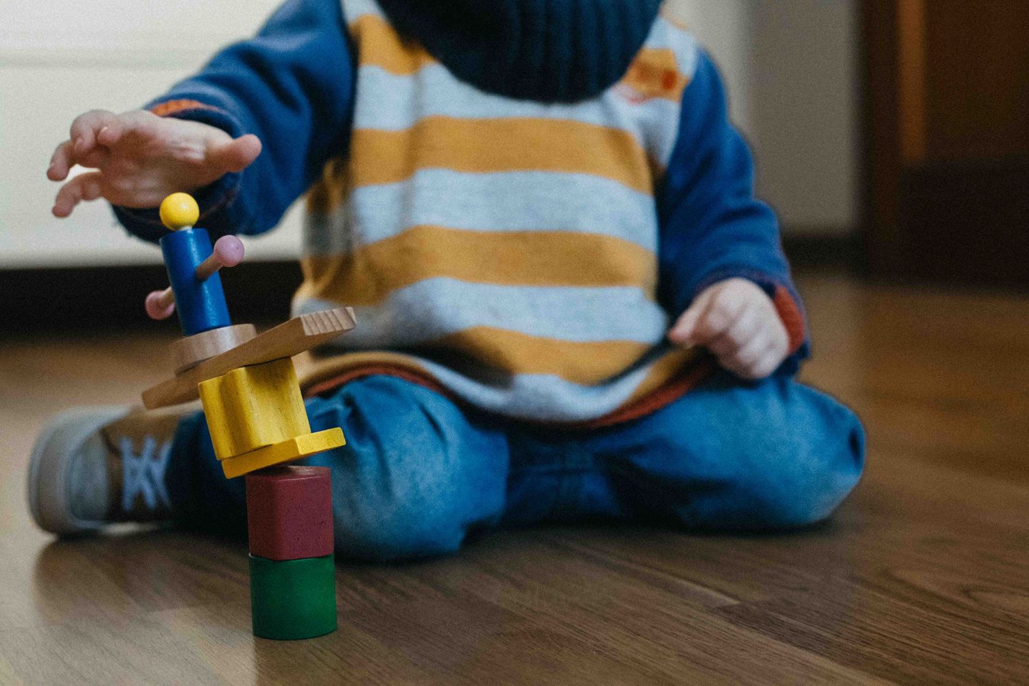 Small child in striped sweater and jeans sits on hardwood floor playing with colorful wooden toy blocks and stacking pieces.