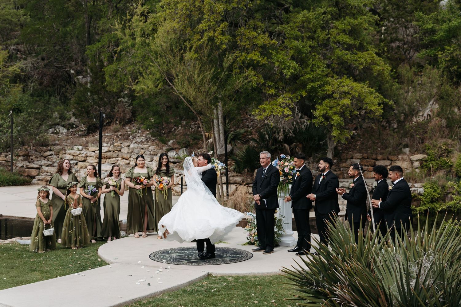 Bride and groom share first kiss at outdoor wedding ceremony with bridesmaids in olive gowns and groomsmen nearby.