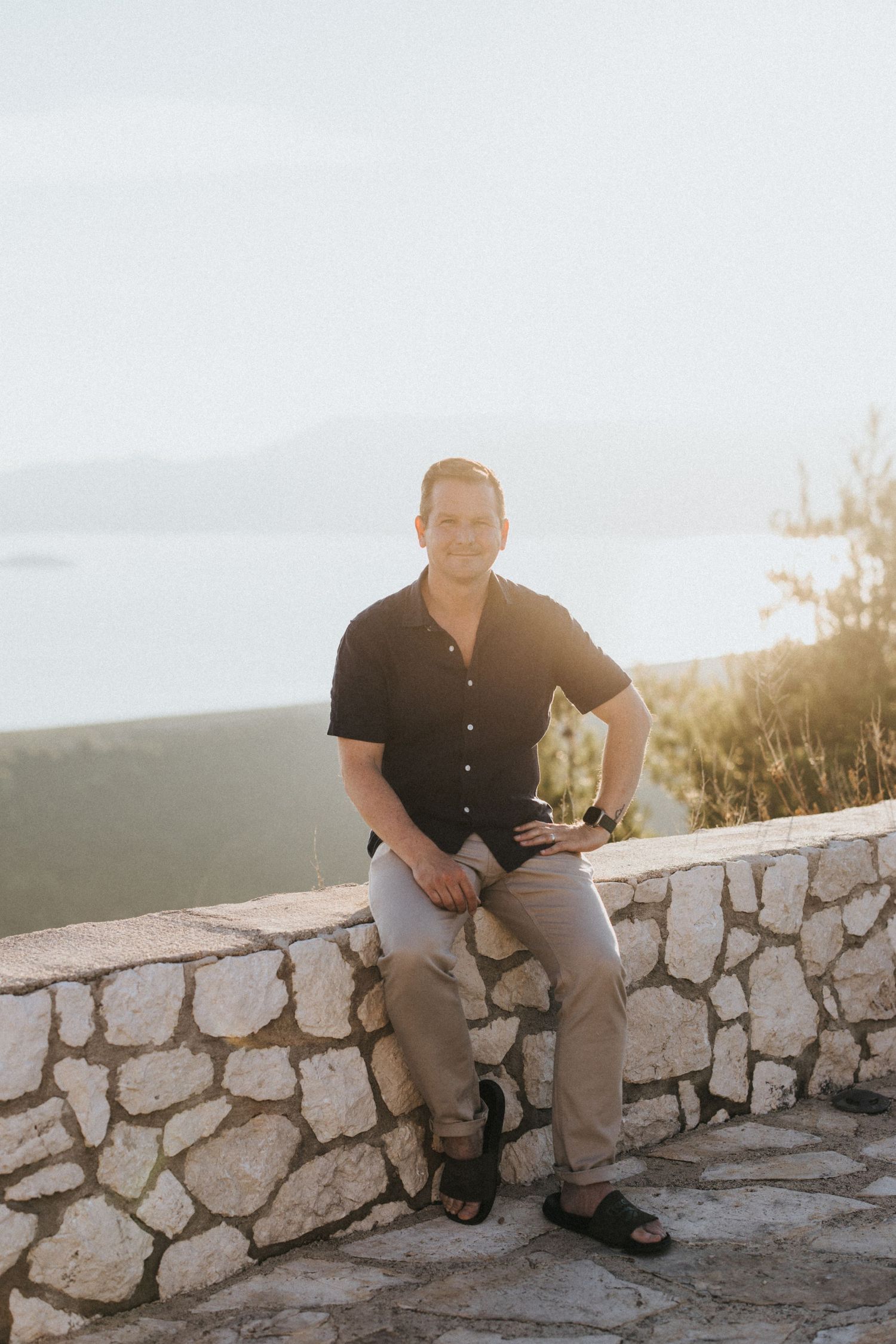 An image of James from James Green Studio, sitting on a stone wall overlooking a scenic mountain vista at sunset.