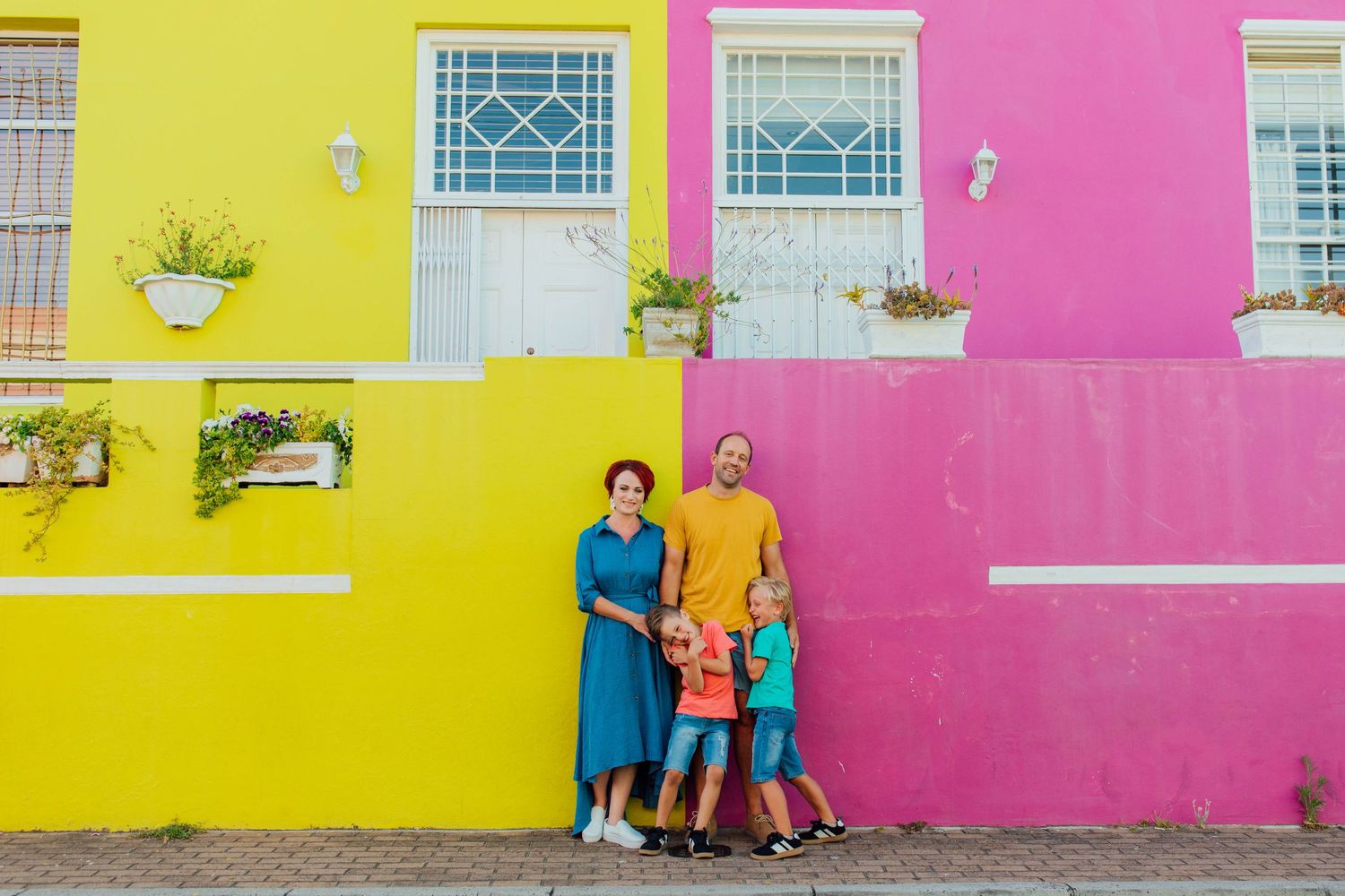 A family standing in front of a colorful yellow and pink building walls with decorative windows and potted plants in Bo-Kaap, Cape Town.