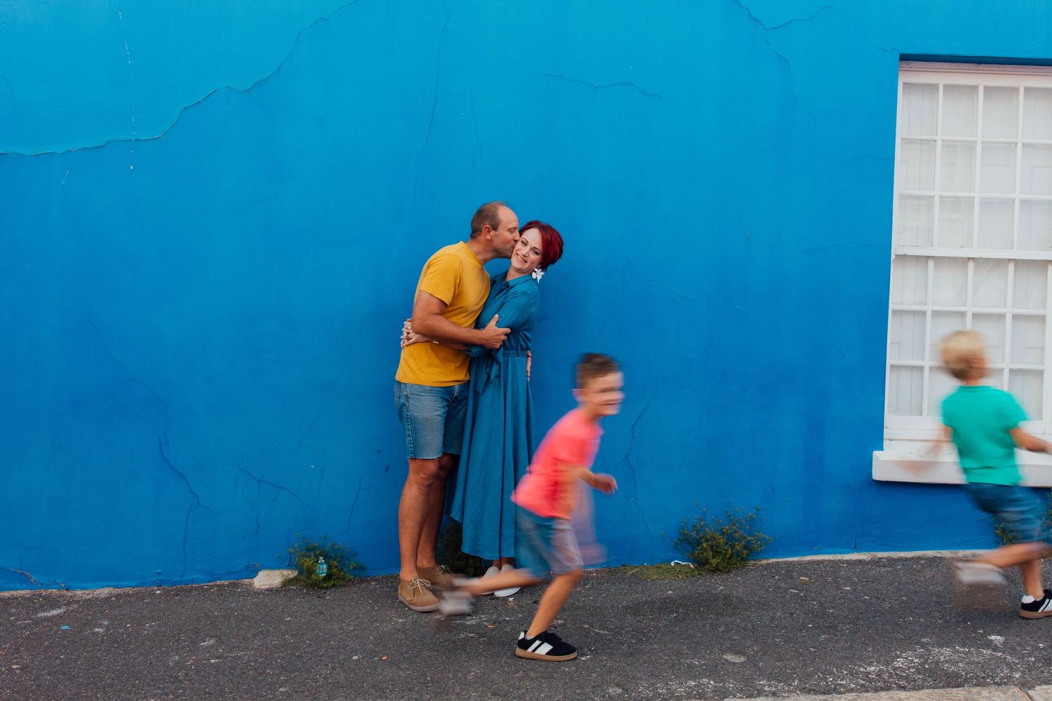 Children play and run past someone photographing against a bright blue wall on an urban street.