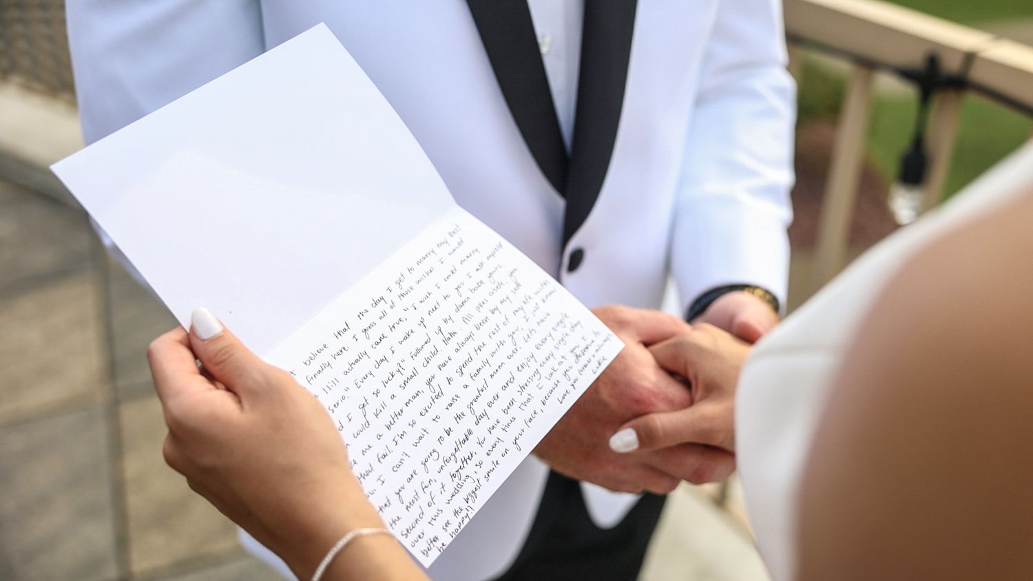 Close-up view of hands holding and reading a handwritten letter during what appears to be a wedding ceremony outdoors.