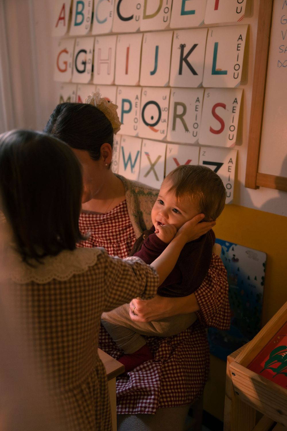 Teacher sits with two young children in a colorful classroom with alphabet letters displayed on the wall.