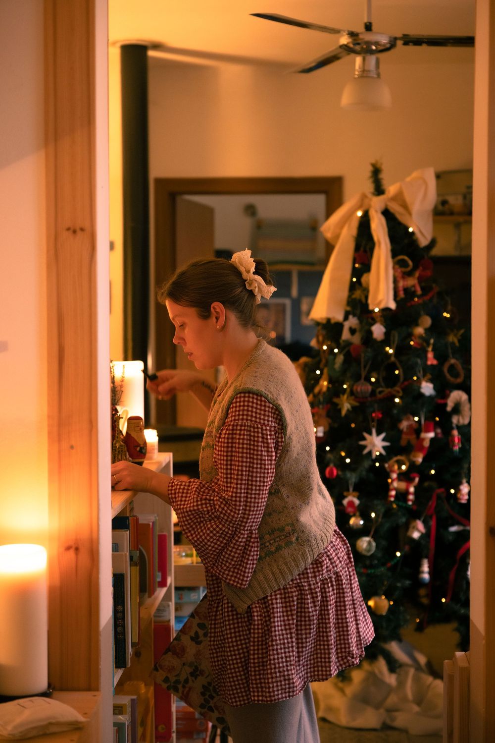 Woman decorating near a candlelit bookshelf with a festive Christmas tree glowing in the background.