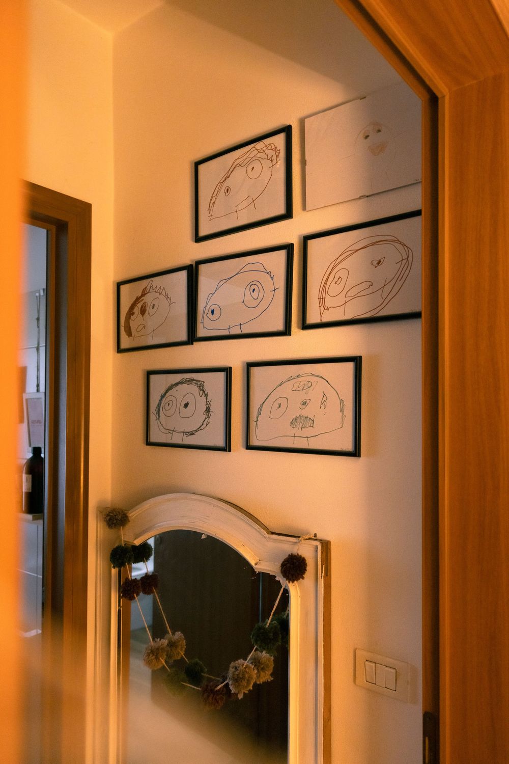 Hallway with framed children's drawings on wall above vintage oval mirror with dried flowers.
