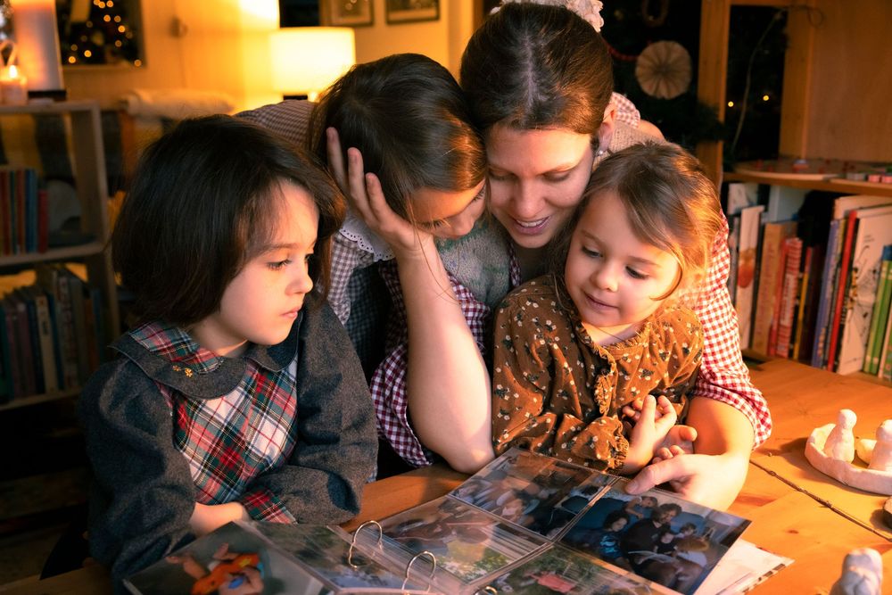 A mother and three children huddle together, looking at a photo album in a cozy, warmly lit room.