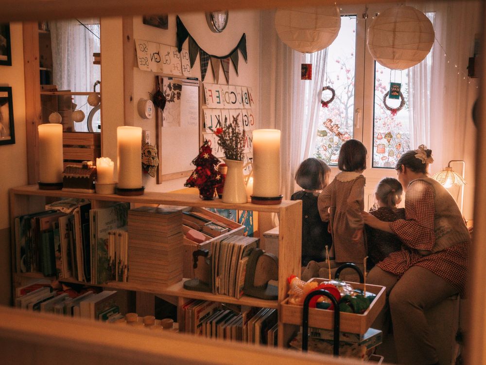 Cozy bookshelf-lined room with lit candles, a mother and child by a bright window, warm holiday decorations.