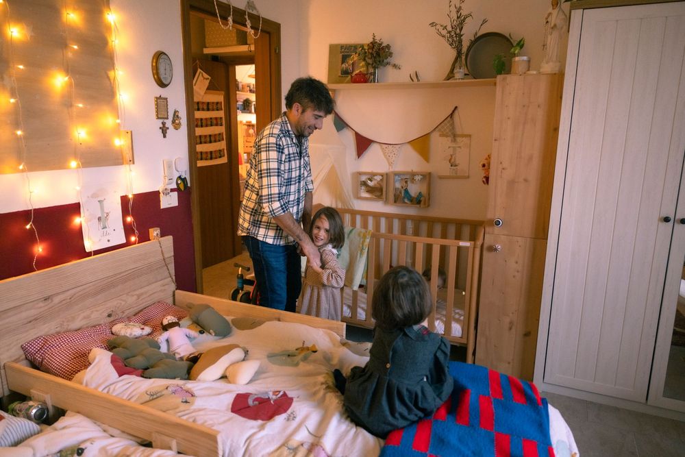 A father and two children play together in a cozy bedroom with warm string lights and a wooden crib.