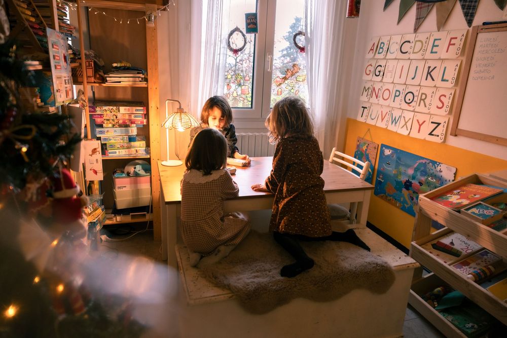 Two children sit at a desk near a window in a cozy room with alphabet chart and Christmas tree decorations.