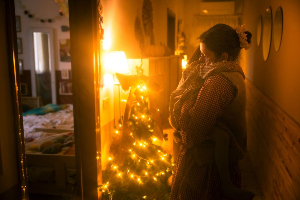 Person standing by a glowing Christmas tree with warm fairy lights in a cozy home hallway at night.