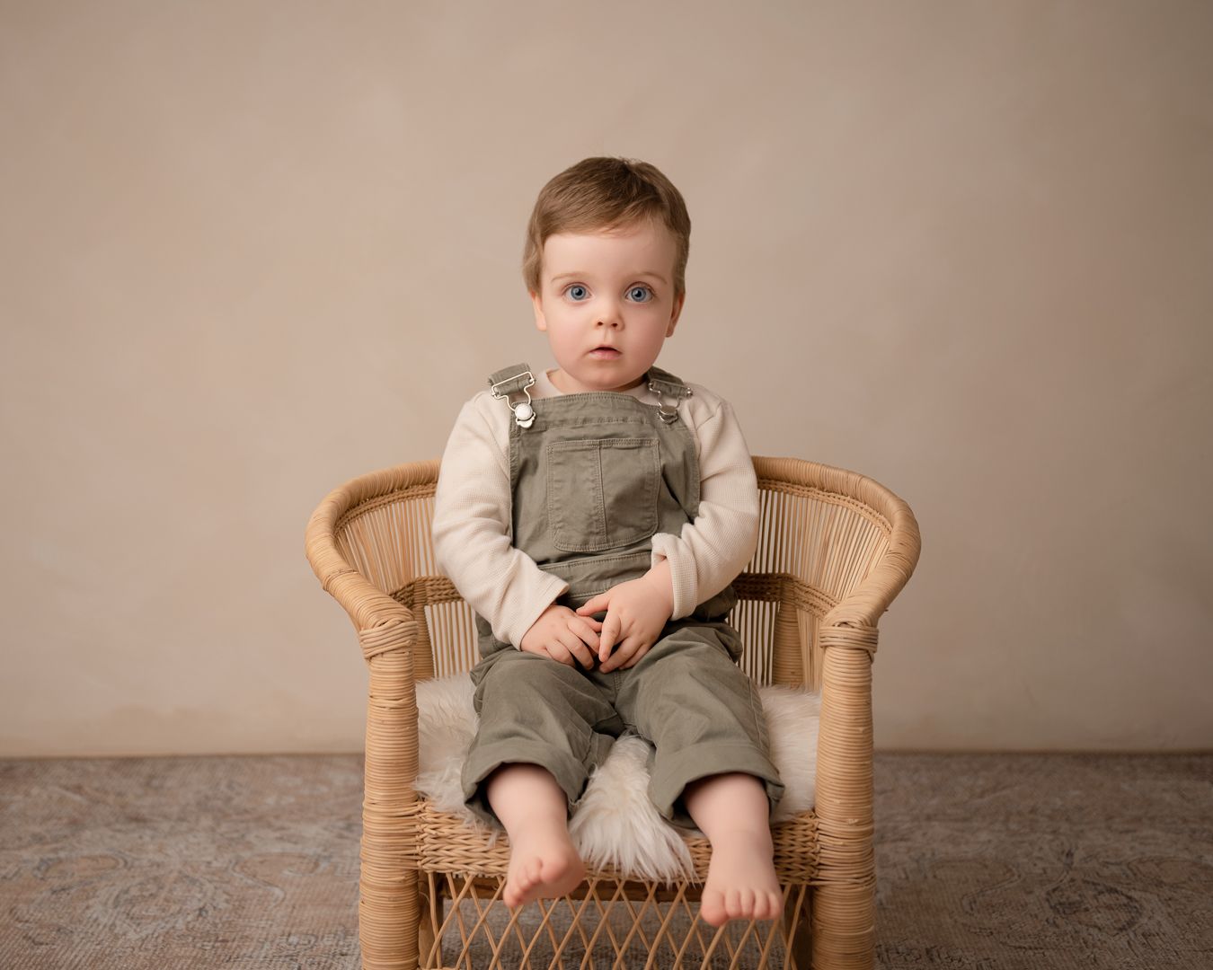 A small child sits in a vintage wicker chair wearing olive green dungarees against a neutral backdrop.