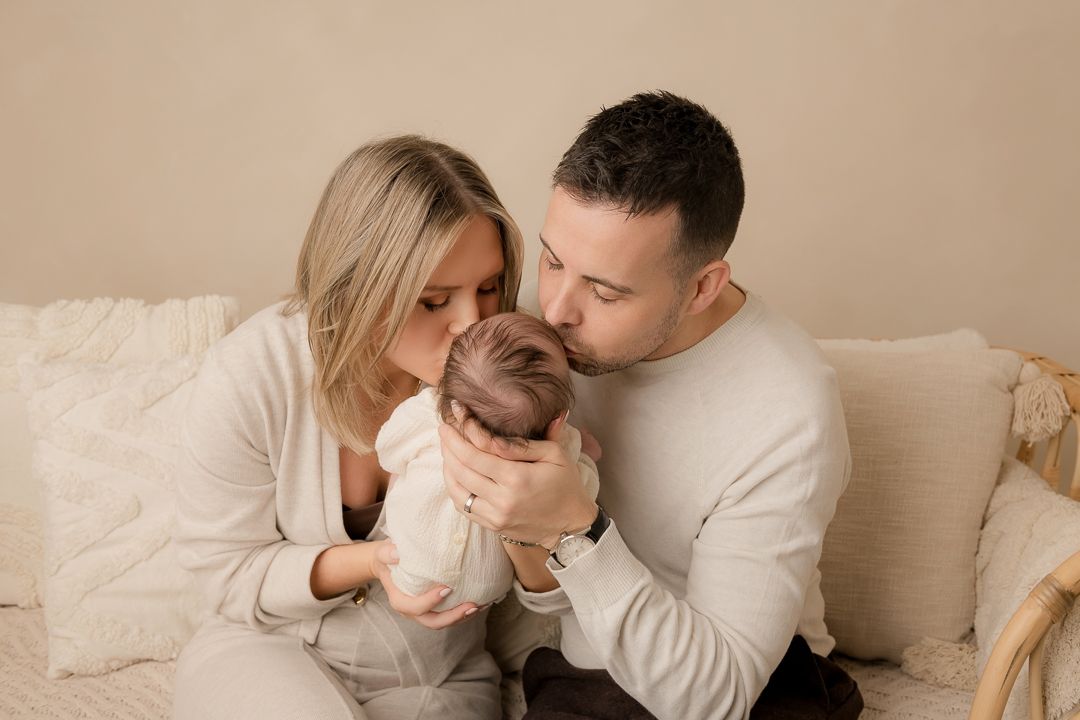 New parents in cream-coloured clothing share an intimate moment with their baby on a couch with neutral-toned lifestyle photography studio.