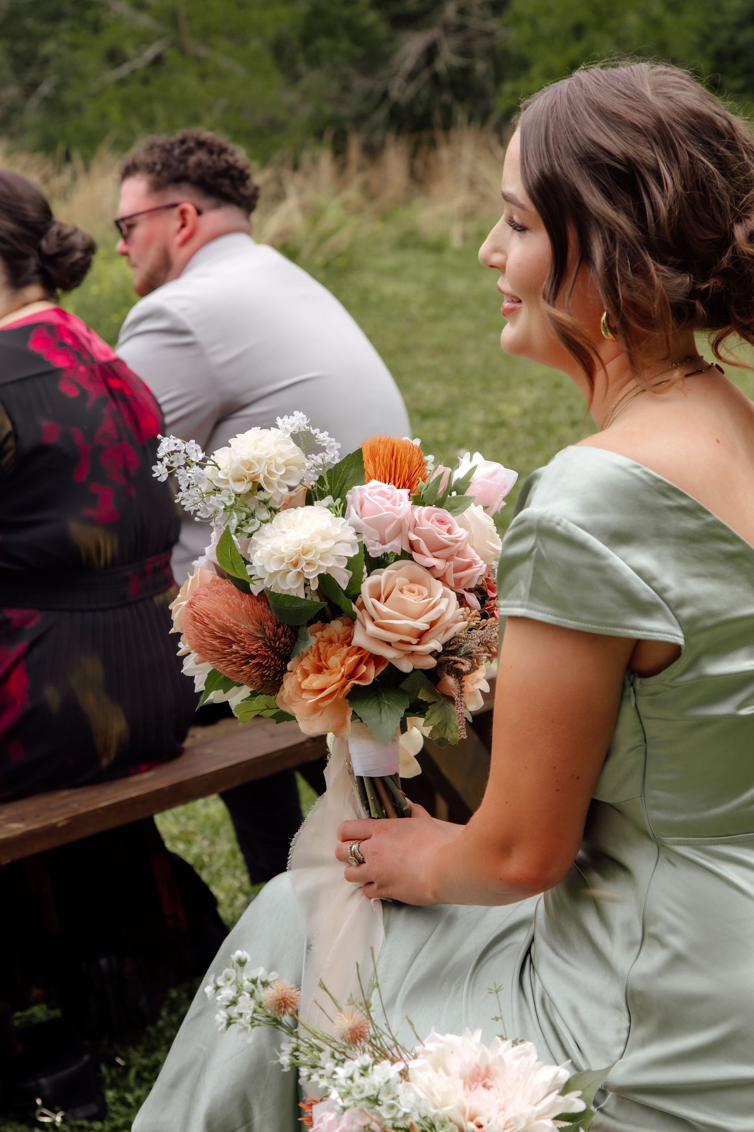 Bridesmaids in sage green dresses hold elegant bouquets of peach and pink roses at an outdoor wedding ceremony.