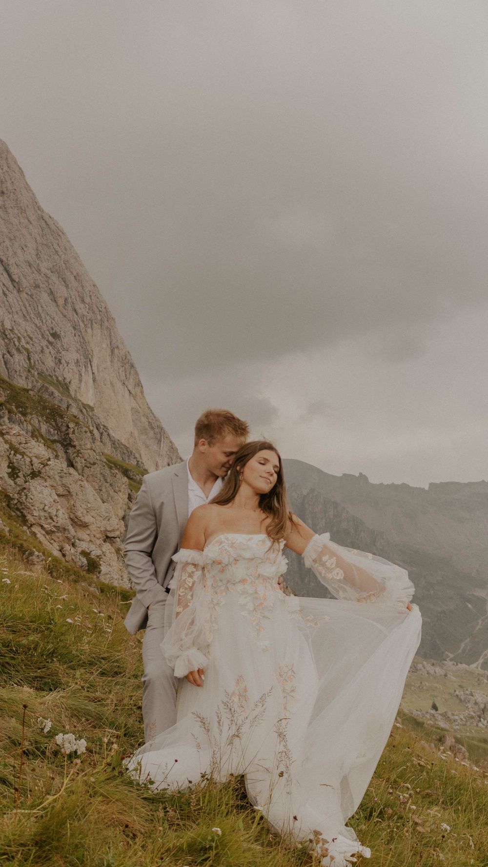 Couple embraces on a misty mountainside with dramatic rocky cliffs and cloudy skies in the background.