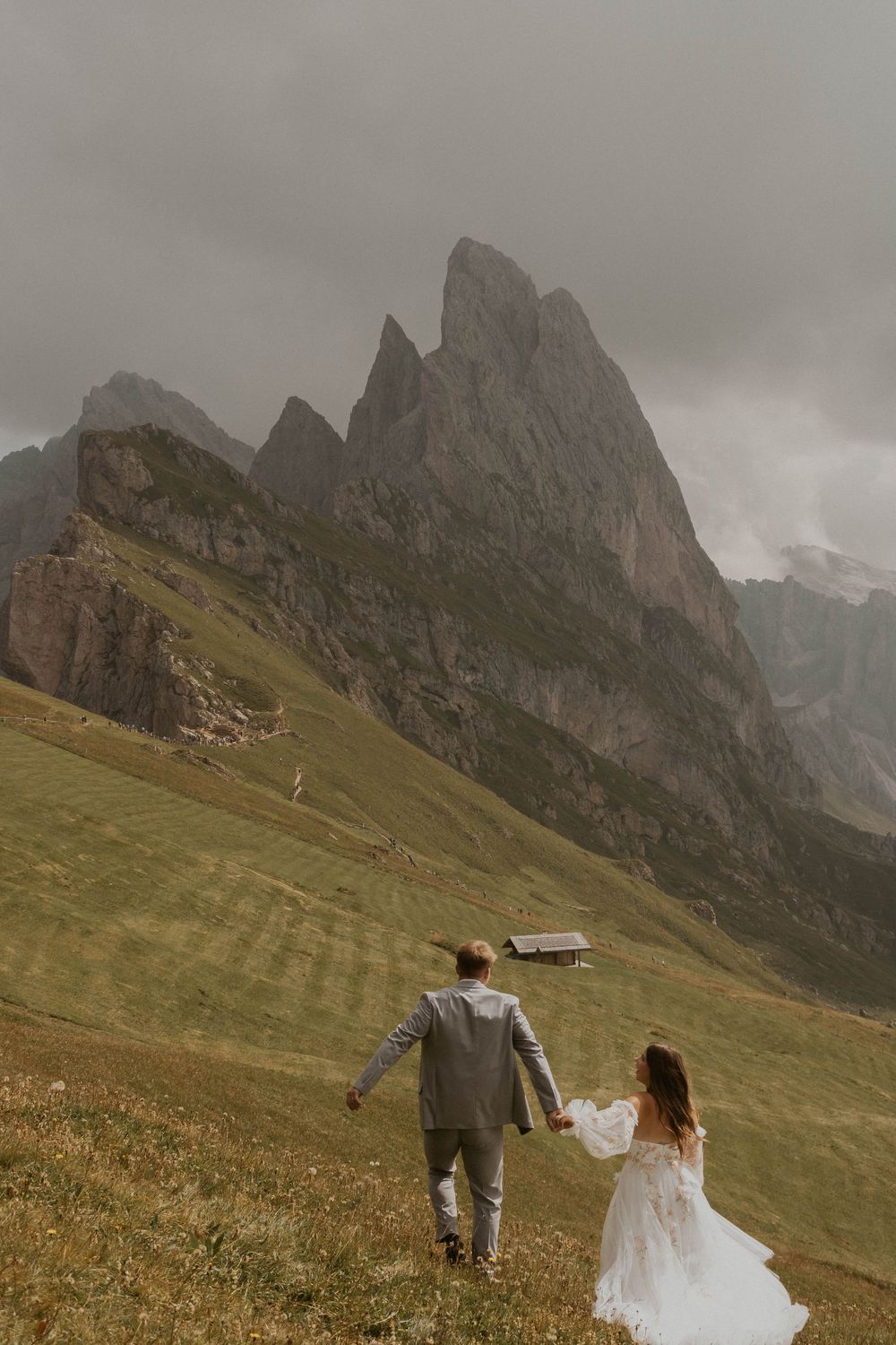 A couple in wedding attire walks across a dramatic mountain meadow in the Dolomites with jagged peaks in the background.