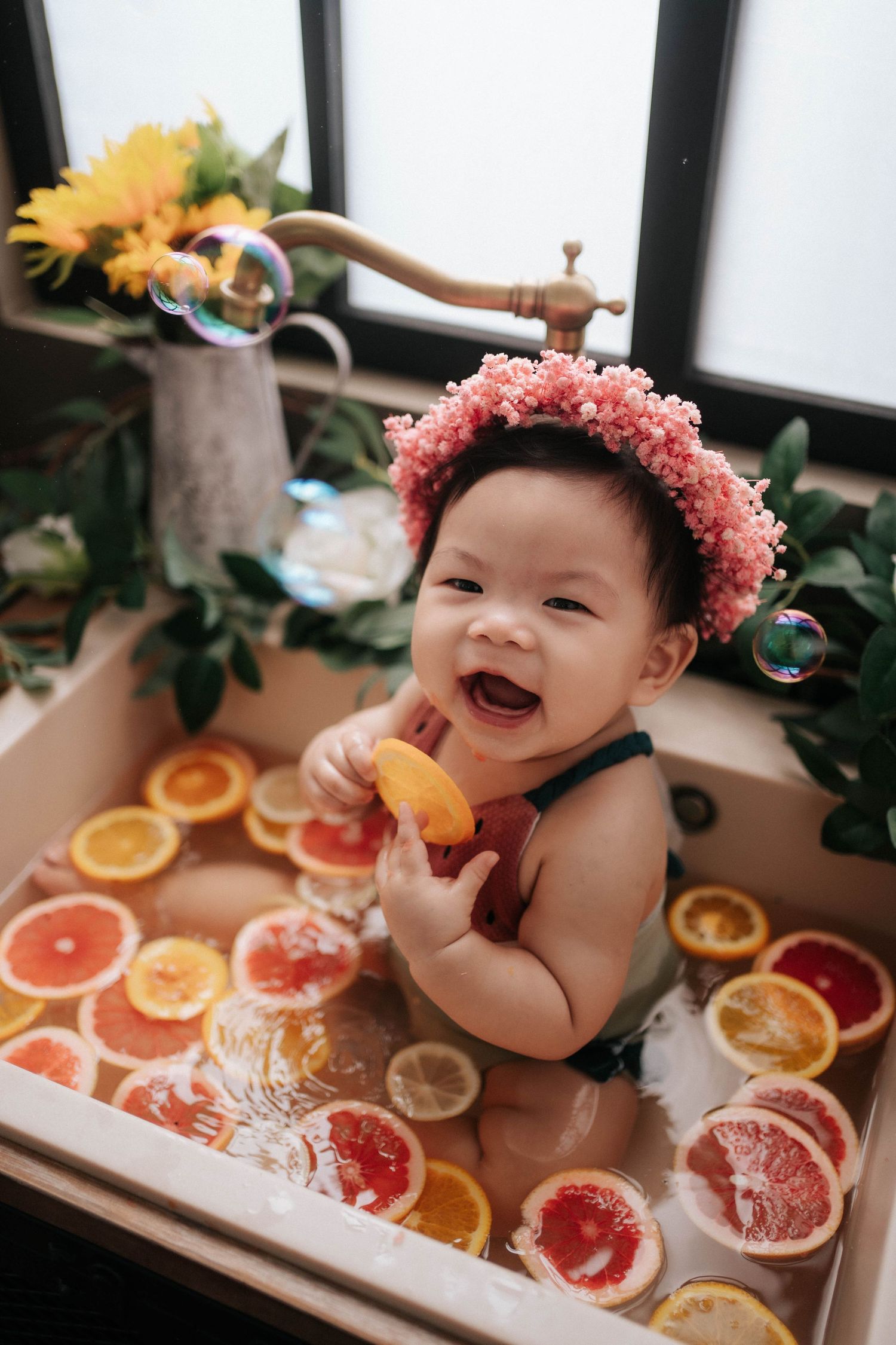 Baby wearing pink flower crown laughs while sitting in bath surrounded by citrus fruit slices.