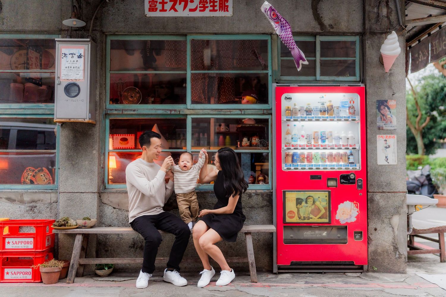 Two people sit on a bench outside a Japanese storefront with a bright red vending machine and traditional decor.