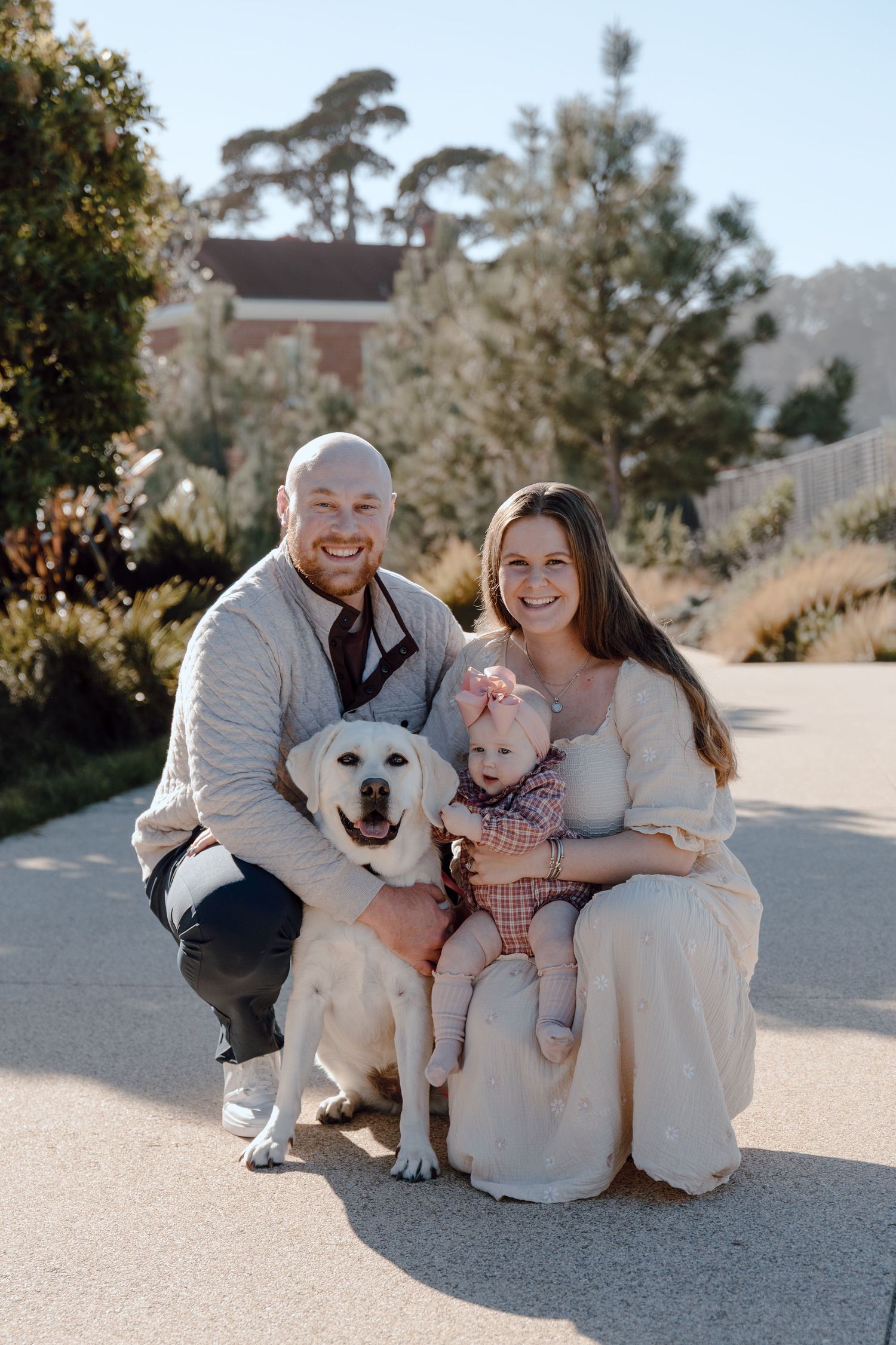 Family sitting together on ground with their white dog in a sunny outdoor setting.