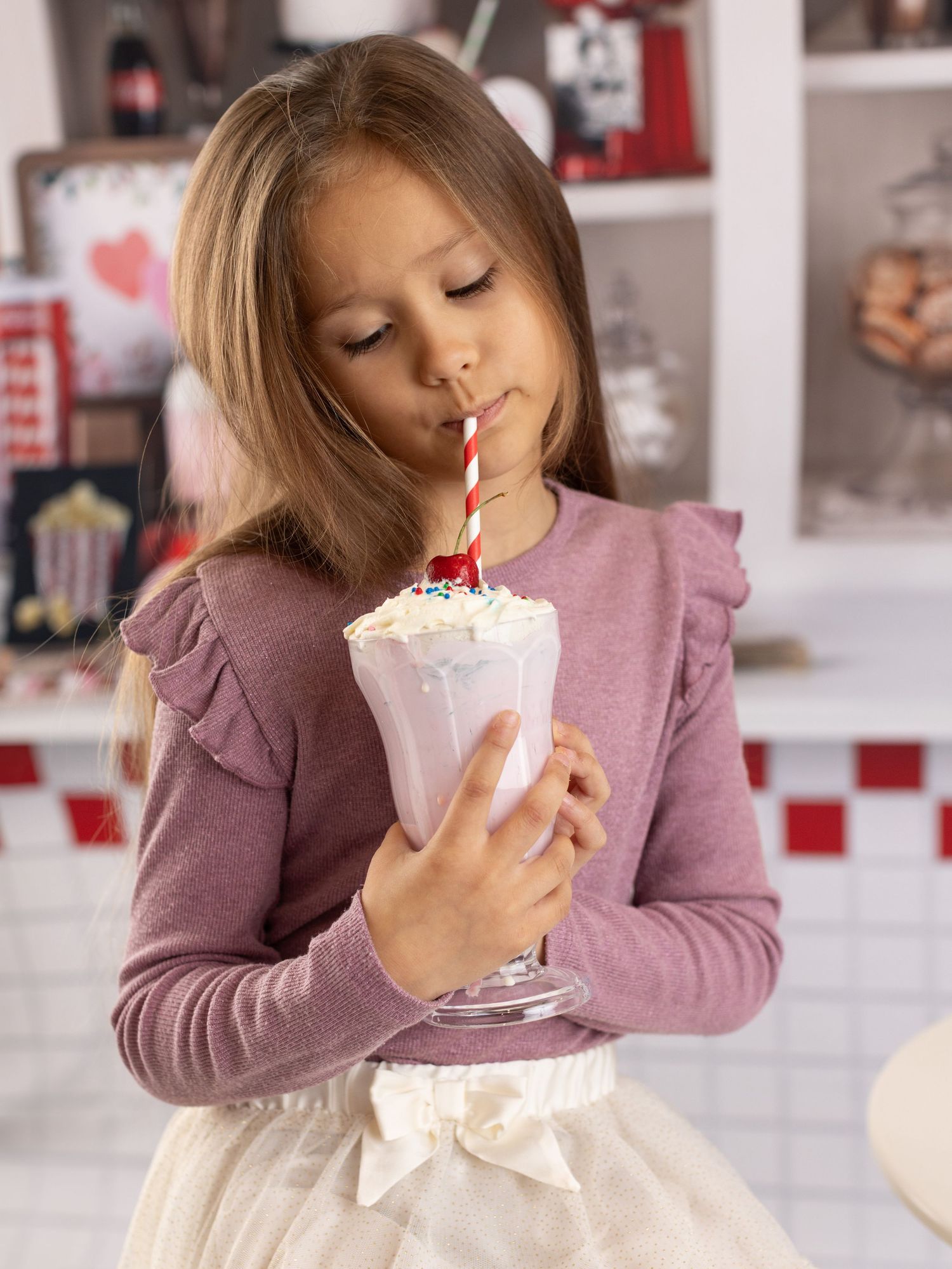 A child in a pink sweater sips a milkshake topped with whipped cream through a red and white striped straw.