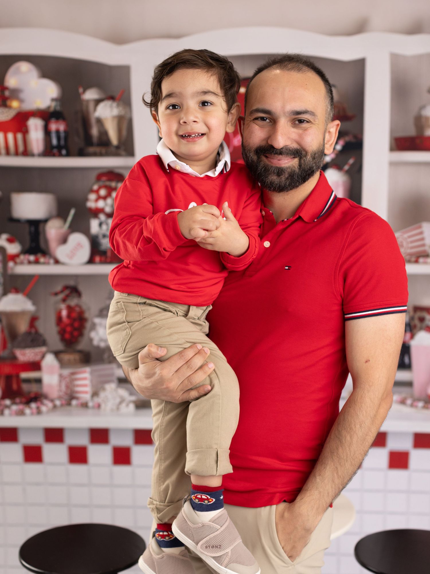 A person in a red shirt holds a child wearing khaki pants in a kitchen with red and white checkered tiles.