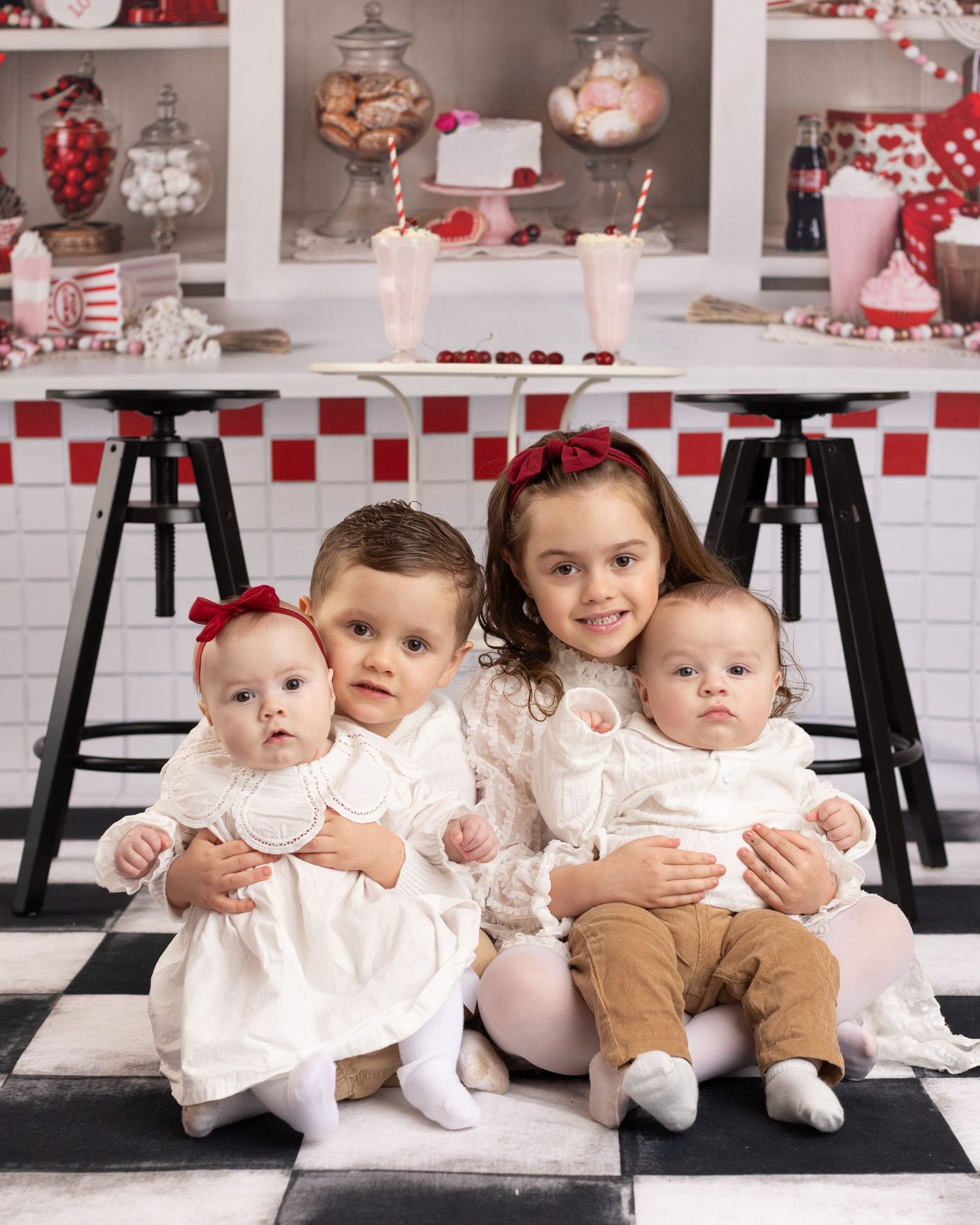 Sweet babies in white dresses sit together on black stools in a festive red and white decorated studio setting.