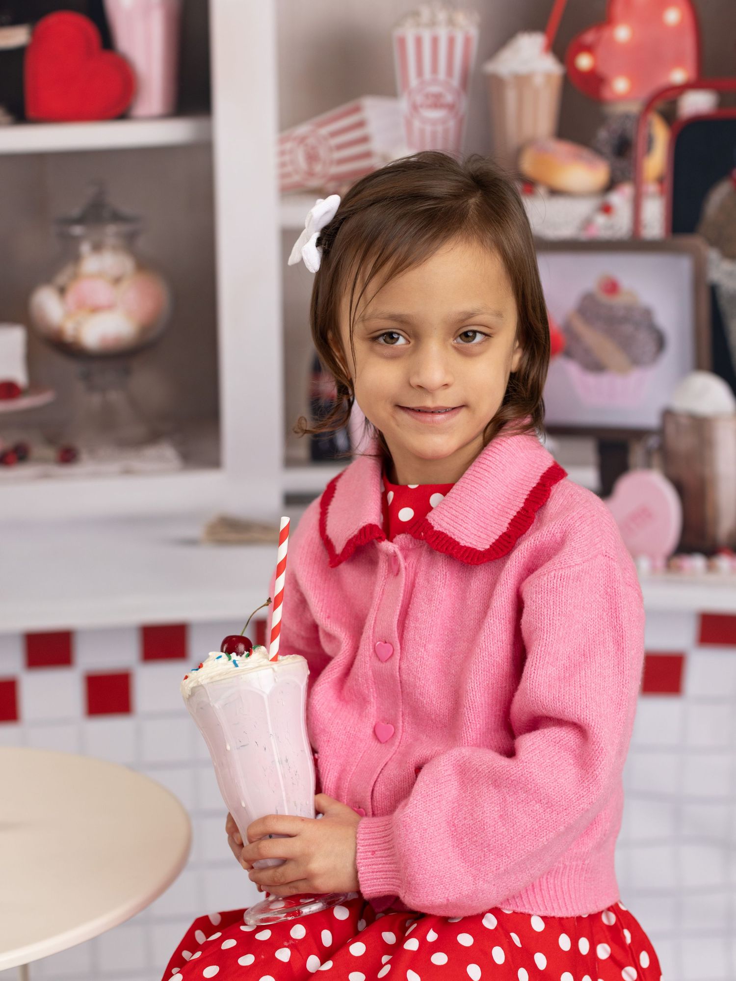 A child in a pink sweater and polka dot dress sits at a table with festive decor in the background.