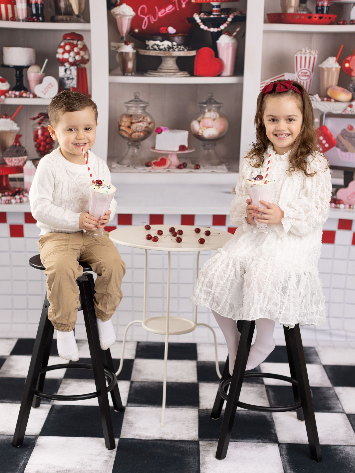 Two children sit on black stools at a vintage-style soda counter with Valentine's decor and red and white checkered floor.