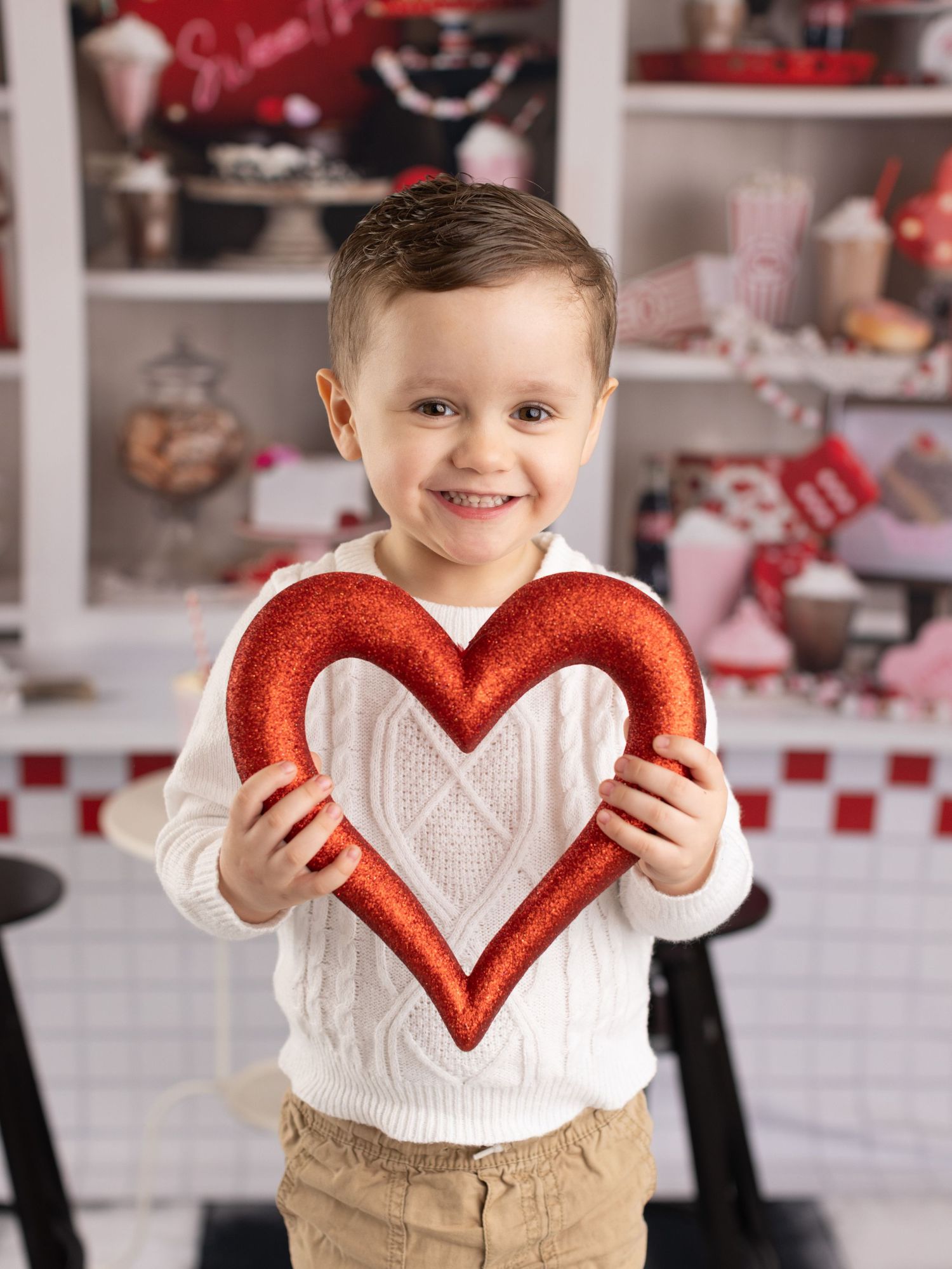 A small child in a white sweater holds a red glittery heart decoration against a festive Valentine's Day backdrop.