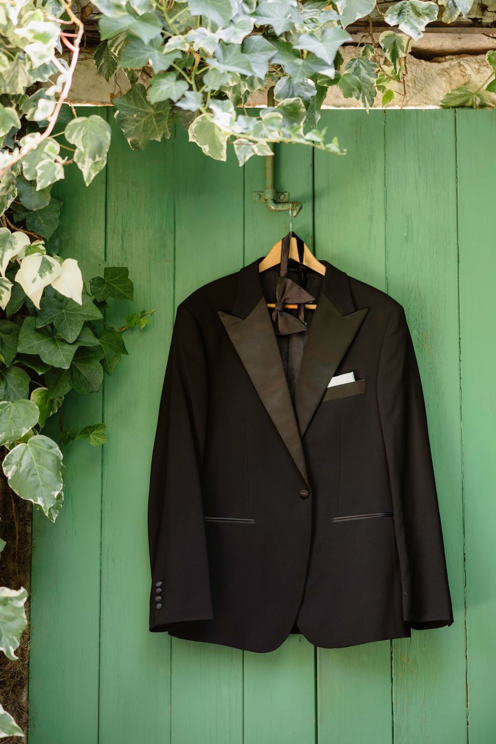 Black formal tuxedo jackets hanging on wooden hangers against a green painted fence with ivy growing above. Hvar, Croatia