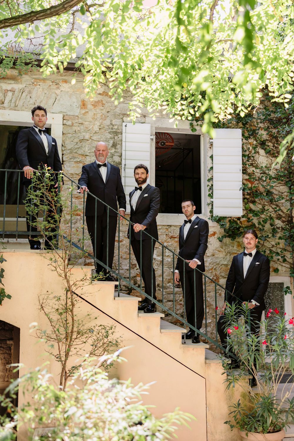 A group in black tuxedos and bow ties pose on elegant stone steps surrounded by lush greenery and white flowers. Hvar, Croatia