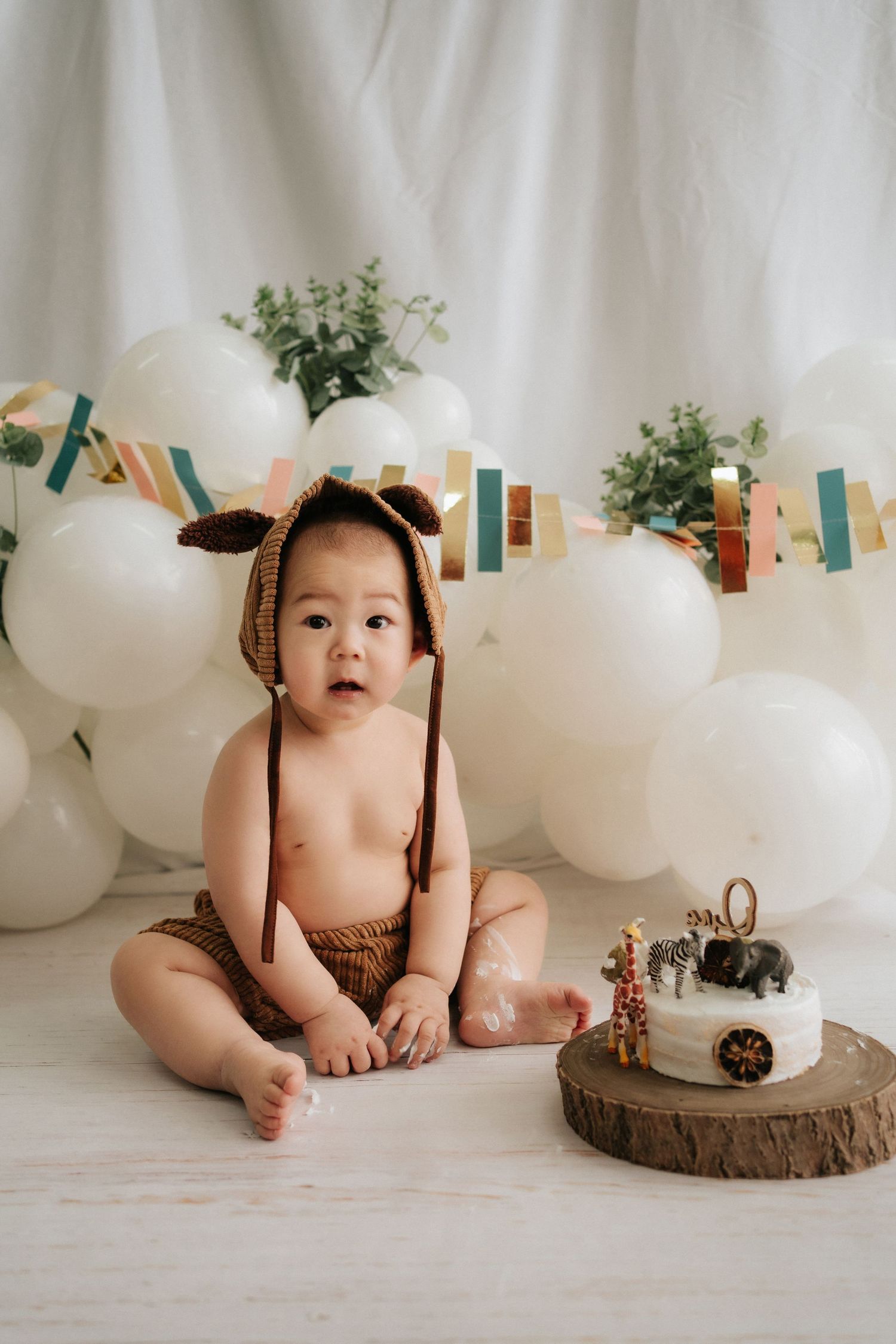 A baby in brown knit outfit sits among rustic wood slices and white balloons in a minimalist studio setting.
