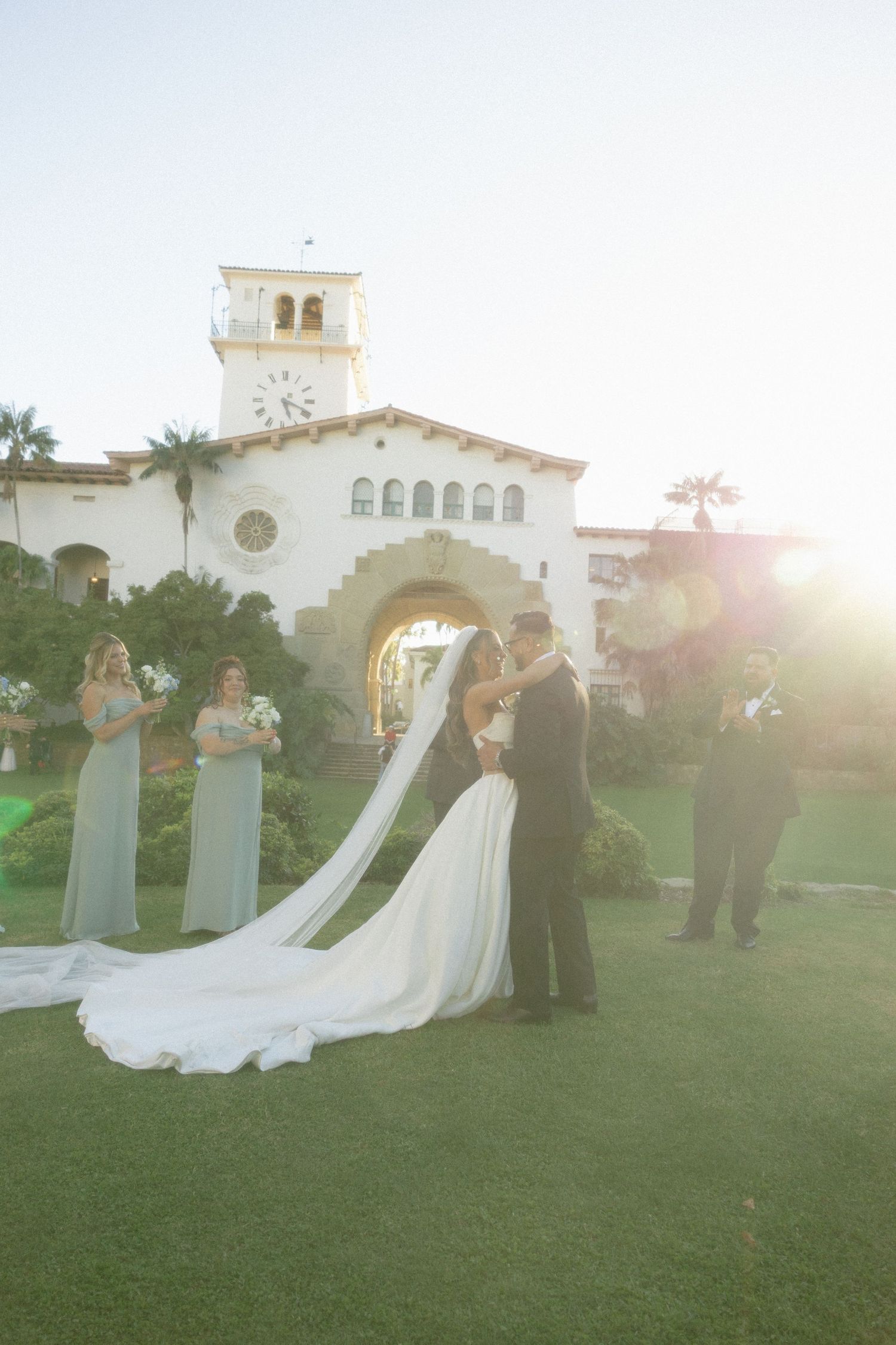 Wedding ceremony taking place on a lush green lawn with Spanish-style architecture in the background at sunset.