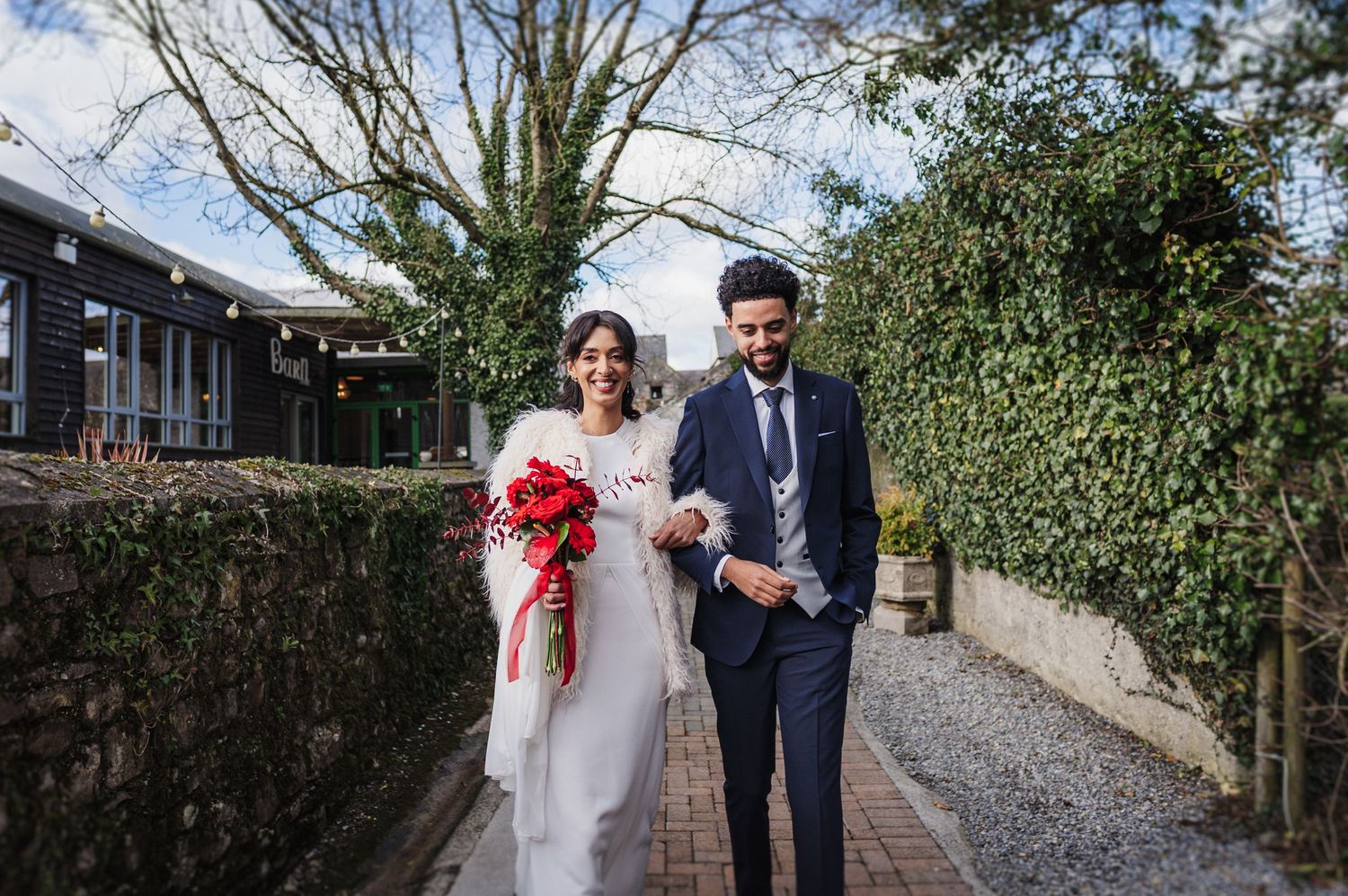 Wedding couple strolling down a tree-lined stone path during an outdoor celebration.