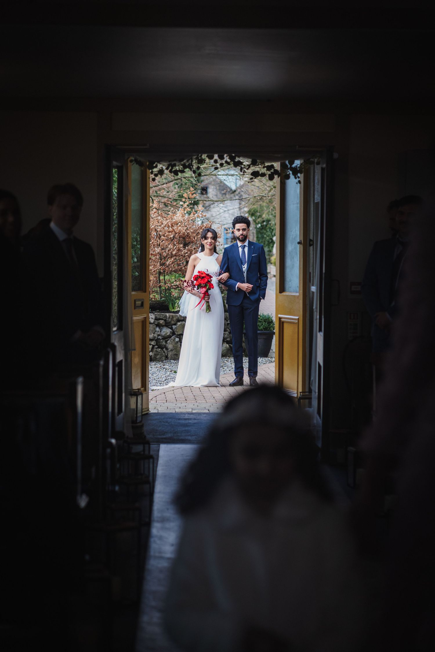 A wedding couple emerges through a dark doorway into bright daylight, creating a dramatic silhouette effect.