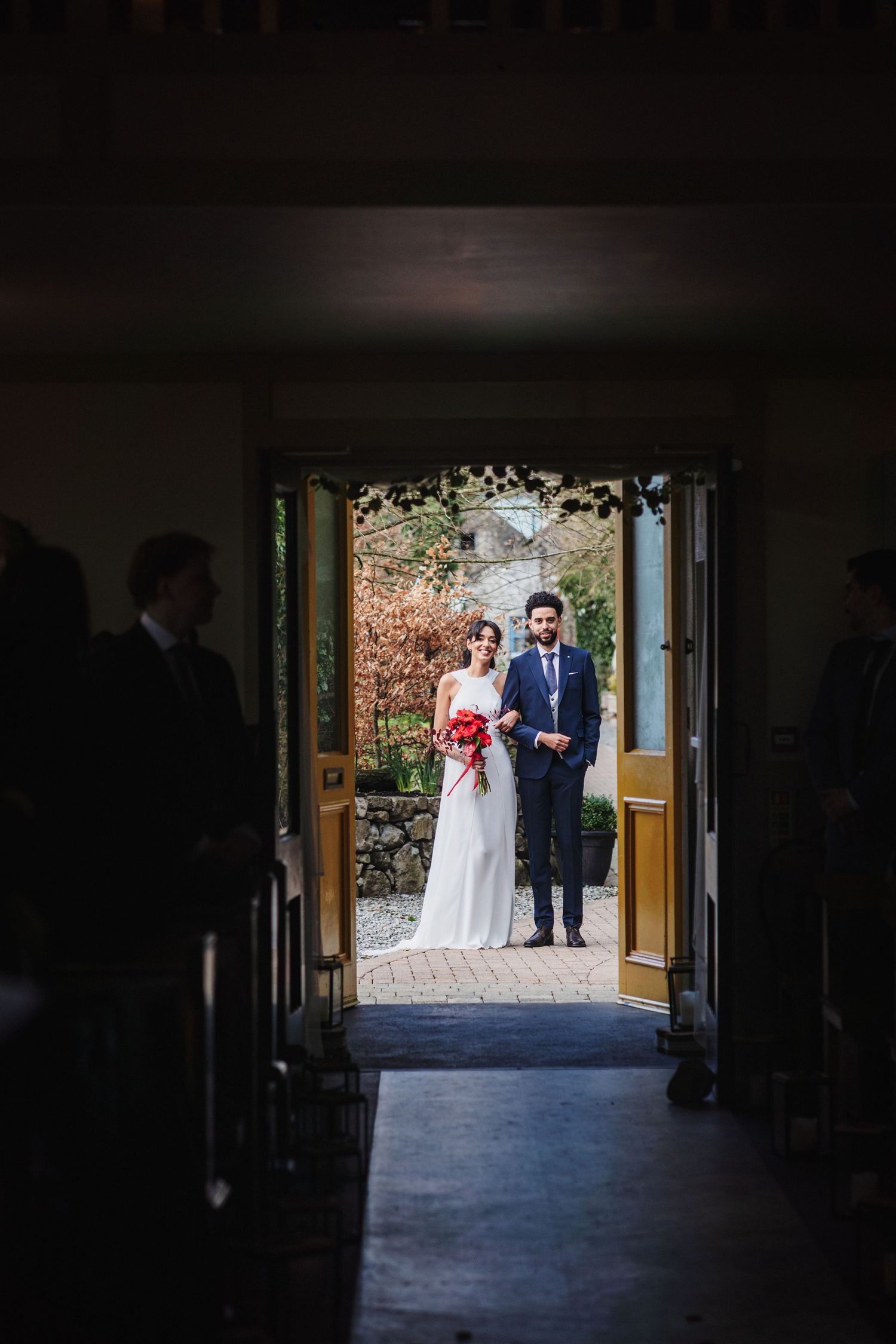 A couple stands framed in a doorway on their wedding day, with autumn foliage visible behind them.