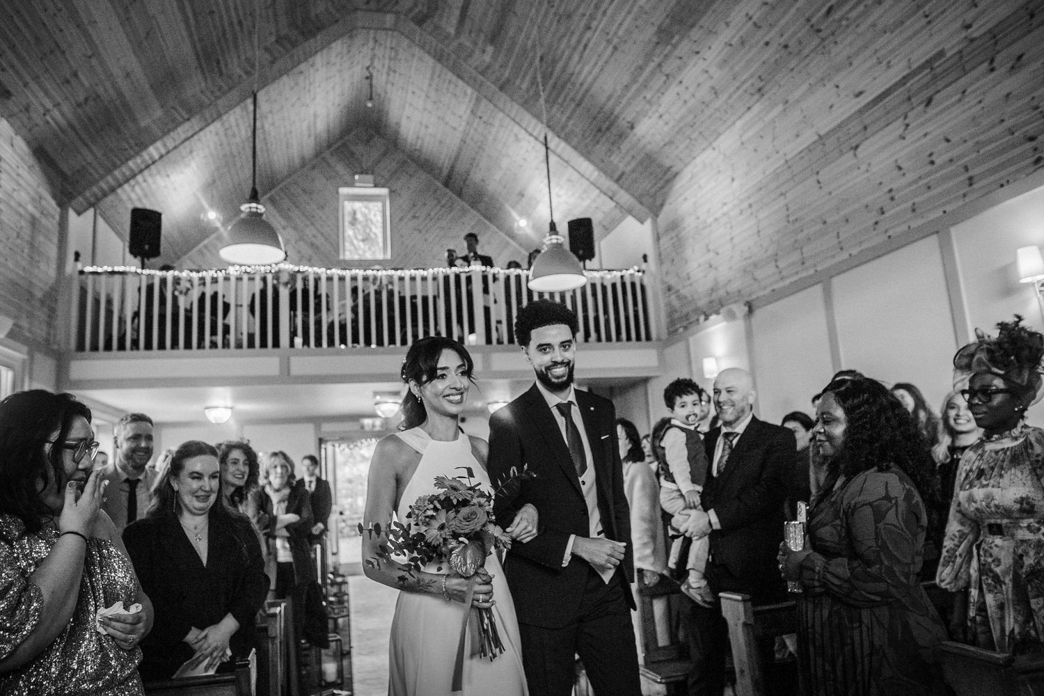 Black and white photo of newlyweds walking down the aisle at a rustic chapel venue with guests and wooden balcony.