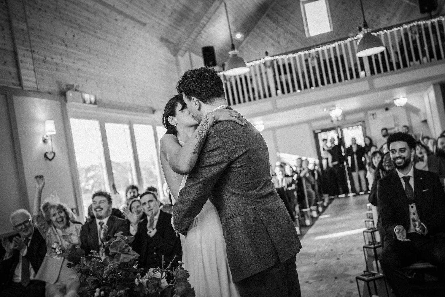 Wedding guests dance and celebrate during a joyful reception in a rustic barn venue with white walls and wooden beams.