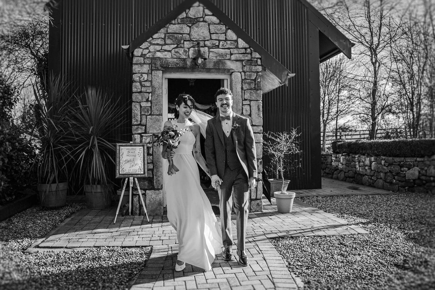 Wedding party poses outdoors in autumn with fallen leaves and rustic barn backdrop in black and white photography.