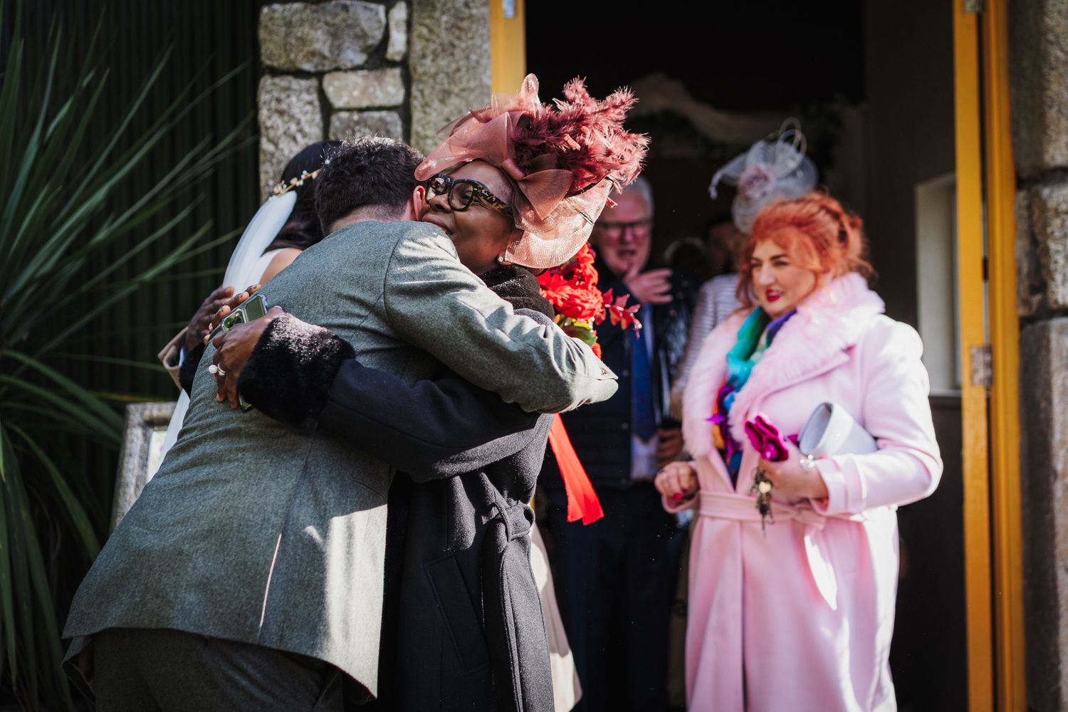 Wedding guests share emotional embraces and greetings outside a stone building with yellow trim.