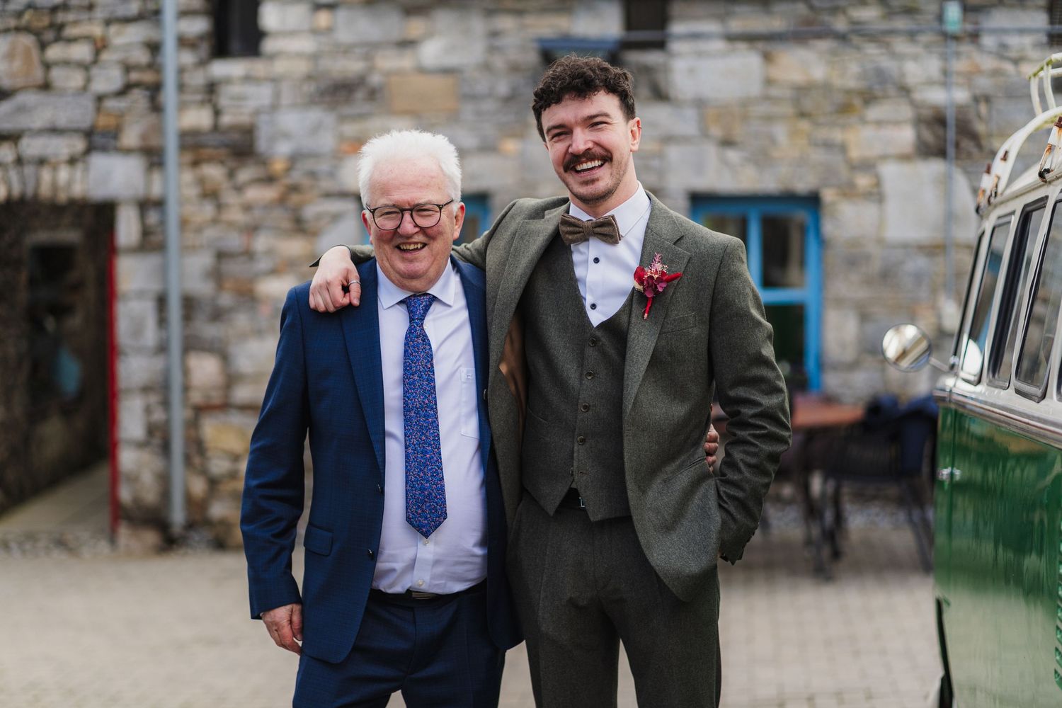 Two people in formal attire share a laugh while walking together outside a stone building.