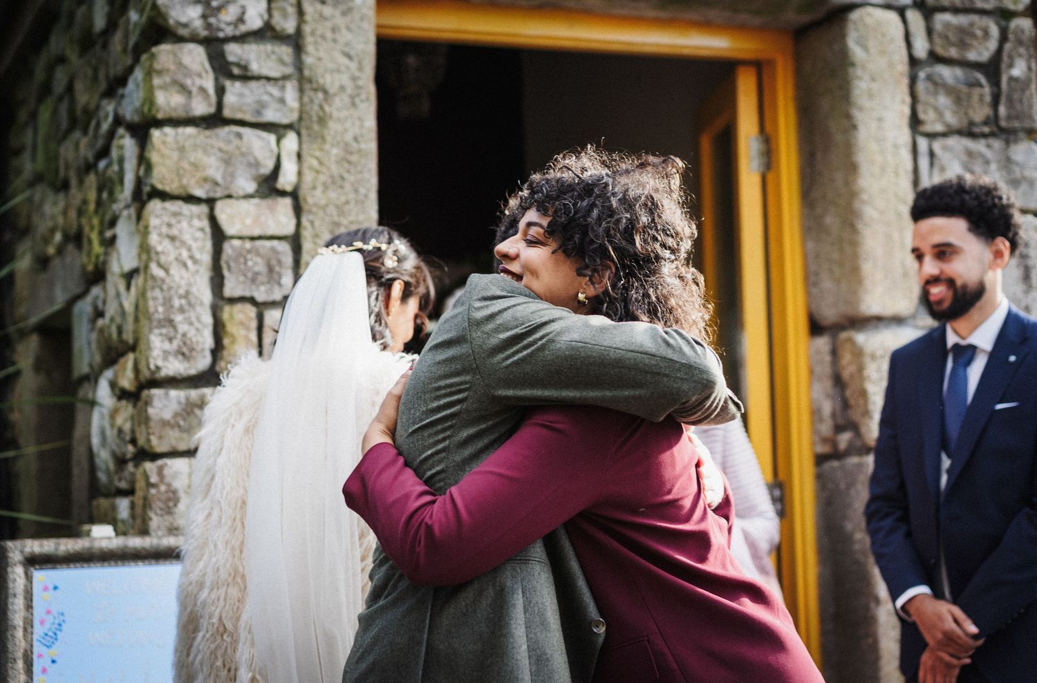 Emotional wedding guests hug and embrace each other outside a rustic stone building entrance.