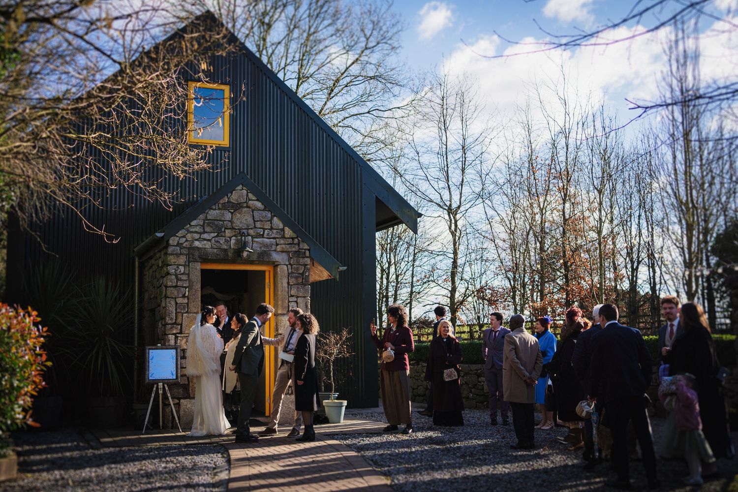 A group gathers outside a rustic stone building on a sunny winter day for an outdoor ceremony.